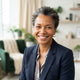 A professional portrait of a smiling, diverse person against a soft-focus background of forest green and warm cream.